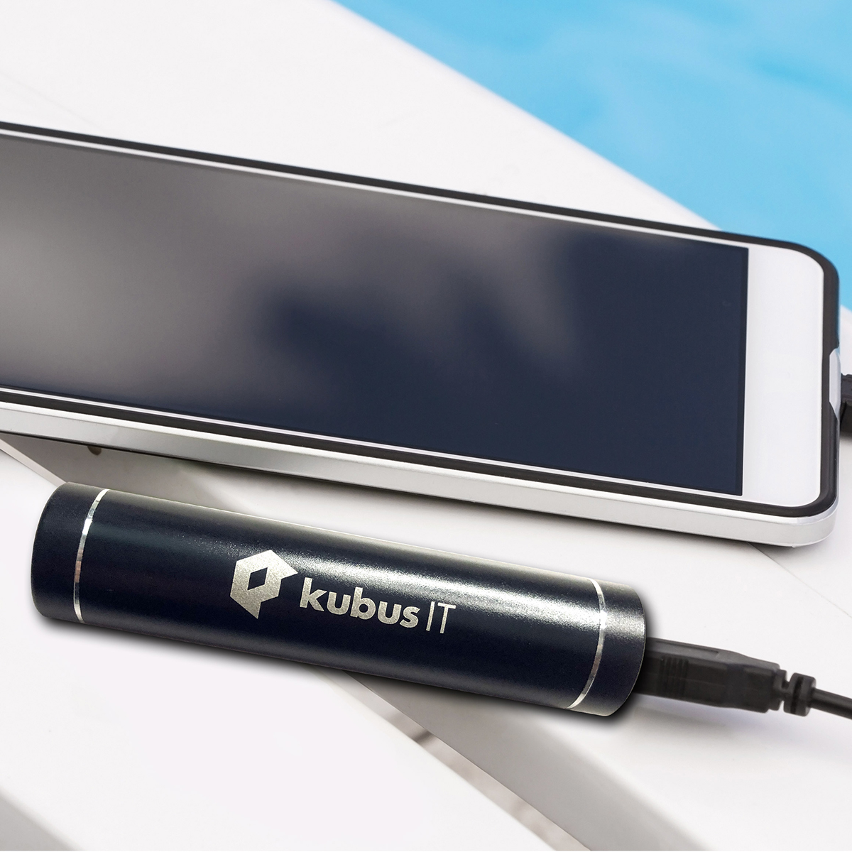 Teaser Quadrat verlinkt, Werbeartikel Powerbank schwarz mit Silberaufdruck kubus IT, liegt neben einem aufzuladenden Mobiltelefon auf einem weißen Stuhl am Poolrand