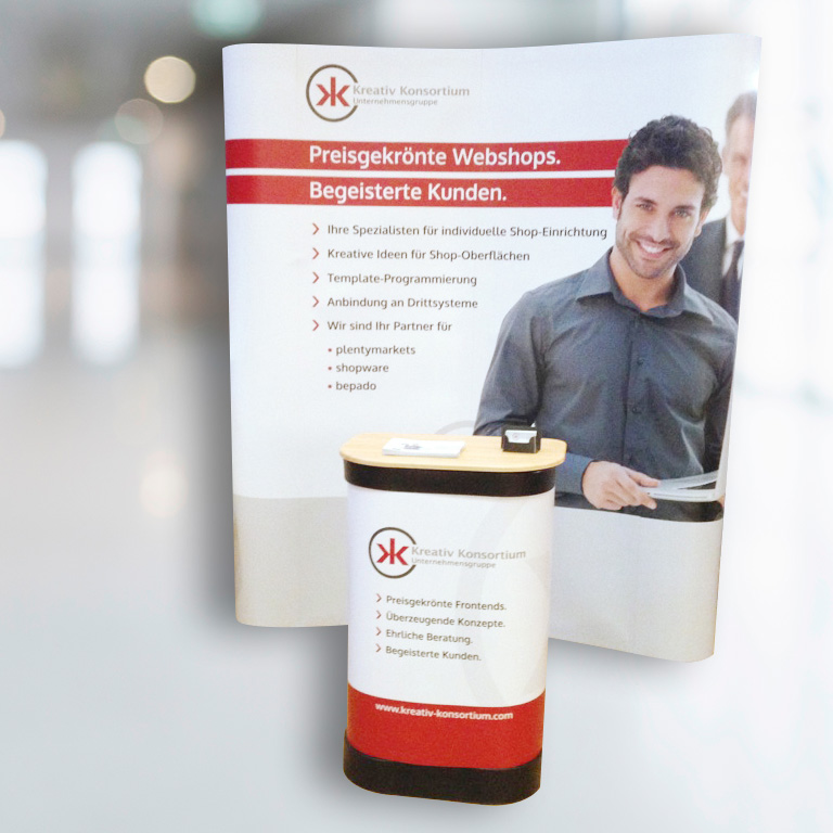 Vorschau Messeausstattung; mobiler Messestand Kreativ Konsortium GmbH, Preisgekrönte Webshops, halbrund mit Logo, Motiv und Text, im Vordergrund ovaler Beratungsdesk; gestaltet mit Logo und Text; Farben Weiß, Hellgrau, Rot